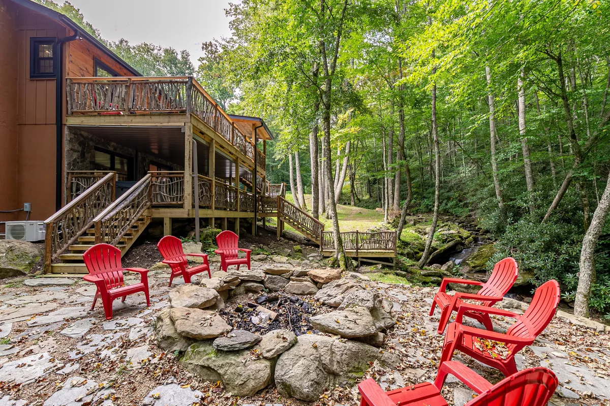 A welcoming scene with a fire pit encircled by red Adirondack chairs on a wooden deck, surrounded by leafy trees. The ambiance suggests communal relaxation—ideal for both summer and autumn outdoor enjoyment.
