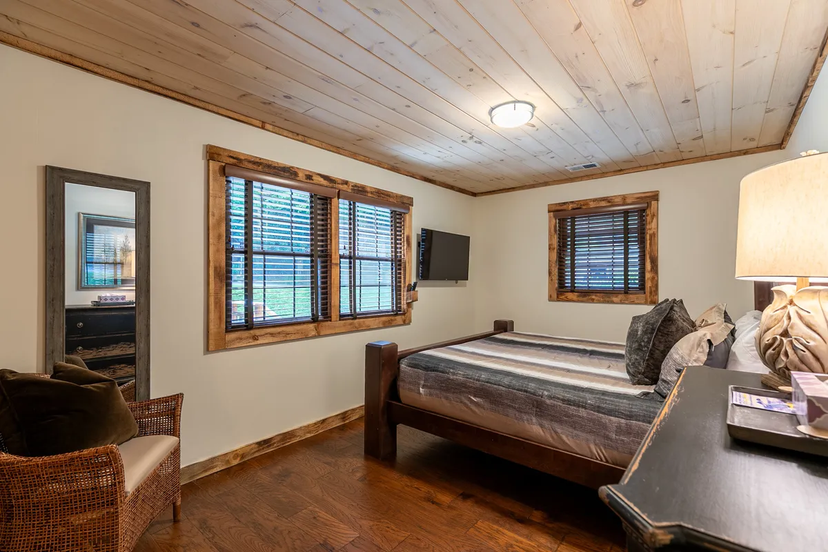 A restful bedroom featuring a neatly made bed with soft, inviting linens. Natural light filters through the windows, illuminating a cozy reading chair, a lamp, and a wall mirror, all contributing to a calm and tranquil atmosphere.