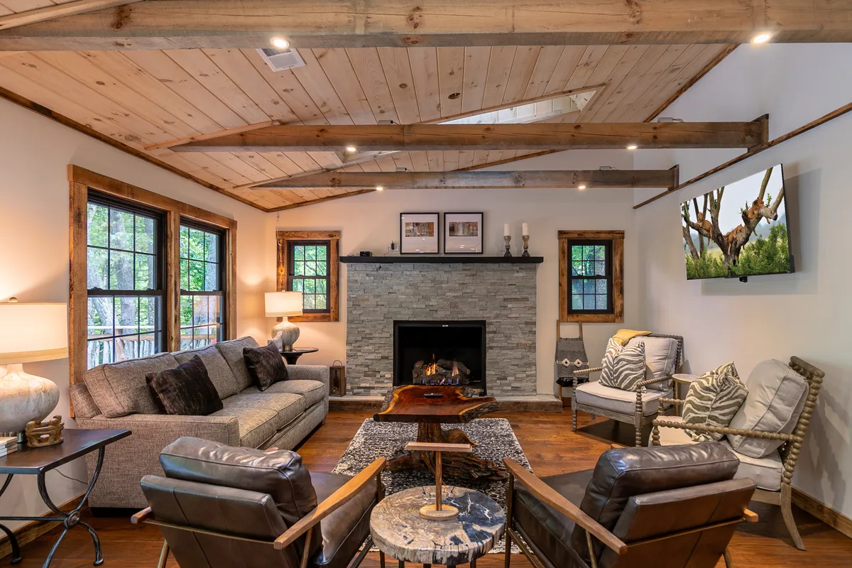 This image captures a spacious living room featuring a striking stone fireplace as the focal point. Comfortable sofas are arranged around the hearth, with wooden beams adding rustic charm to the ceiling. Large windows flood the space with natural light and offer serene views. A television displays a nature scene, and tasteful artwork adorns the walls, creating a warm, inviting atmosphere for relaxation or group gatherings.