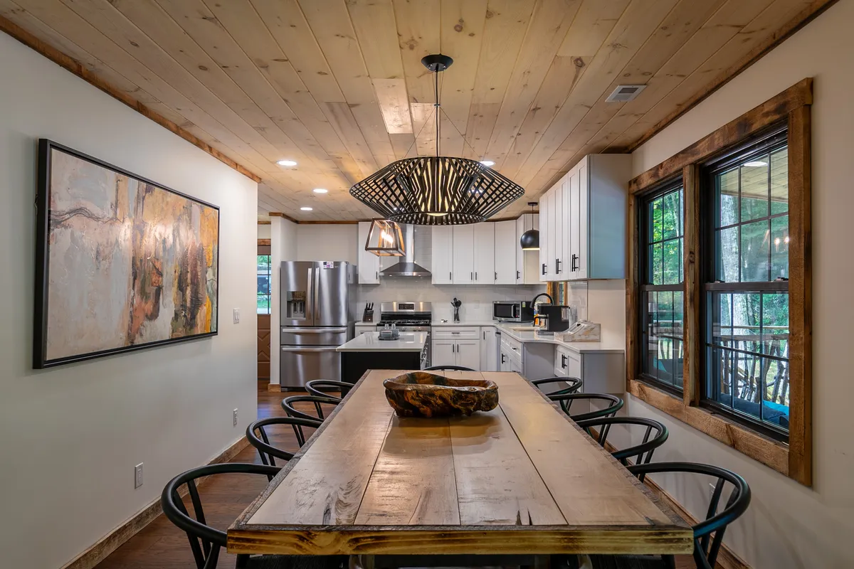 A cozy dining area with a large wooden table, unique light fixture overhead, stainless steel appliances visible in the background, and tasteful wall artwork. The space exudes warmth and is clearly designed for hosting family meals and friendly get-togethers.