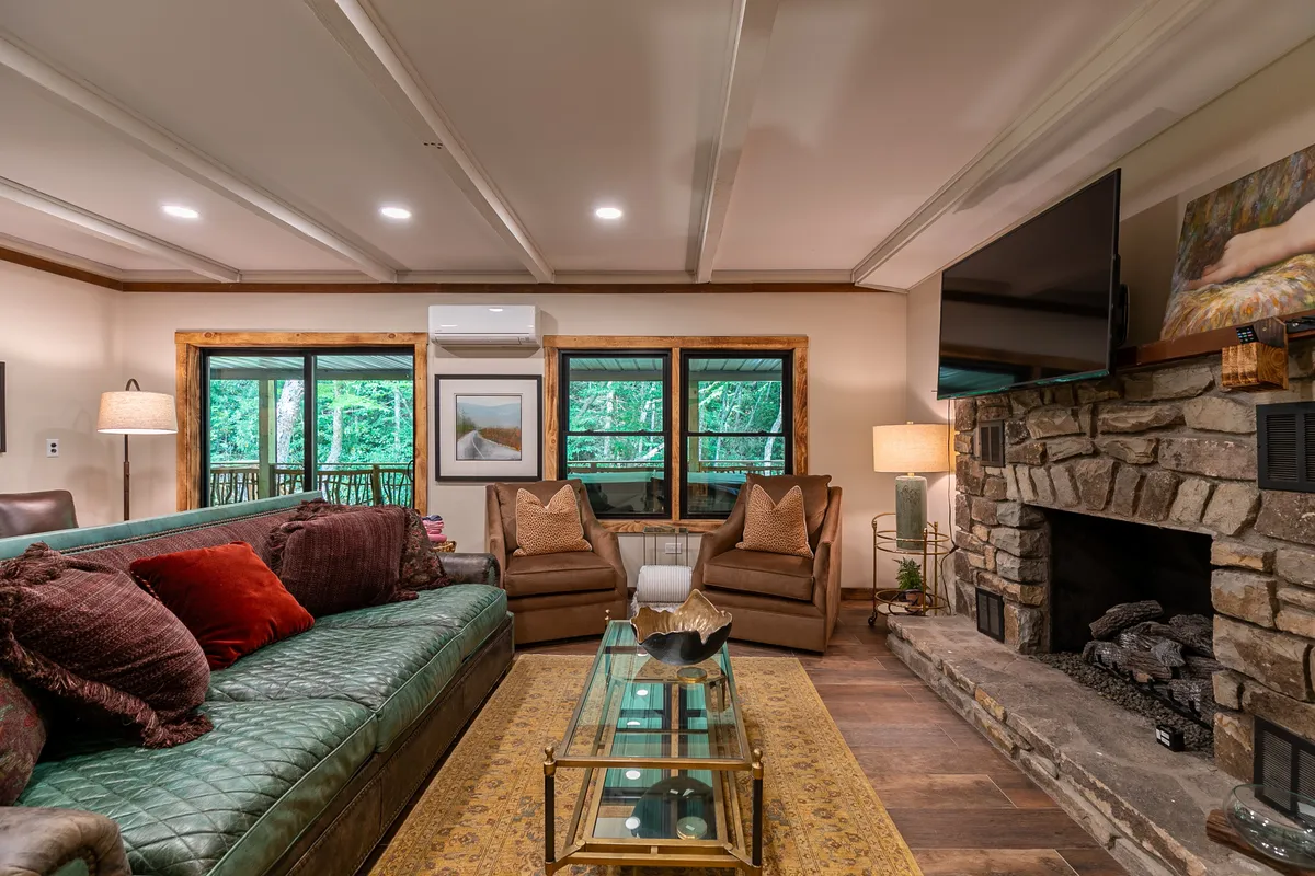 A cozy corner of the main gathering area is highlighted here, featuring a green leather sofa, a classic stone fireplace, expansive windows, and a glass coffee table. The layout encourages conversation and relaxation while offering views of the outdoors, blending comfort with a sense of openness.
