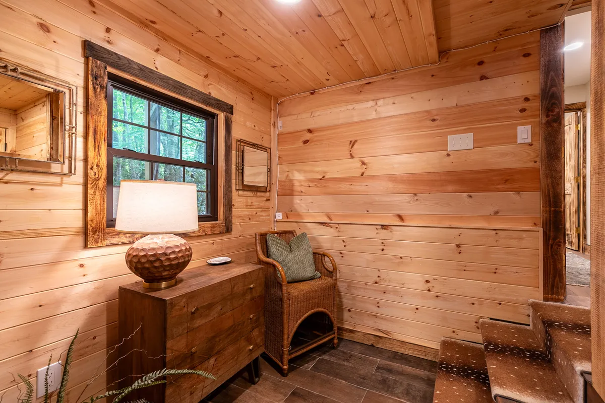 This image displays a warmly lit space with wooden paneling, a lamp, mirror, woven chair, and chest of drawers. The window lets in light and offers a subtle glimpse of the outdoors, evoking the changing scenery beyond.
