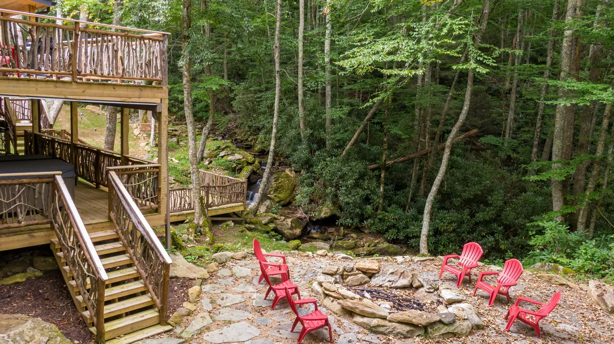 A cozy outdoor setting featuring red Adirondack chairs surrounding a stone fire pit on a wooden deck, all nestled amidst mature trees. The area looks perfect for gathering, storytelling, and relaxing, with the mountain ambiance clearly visible.