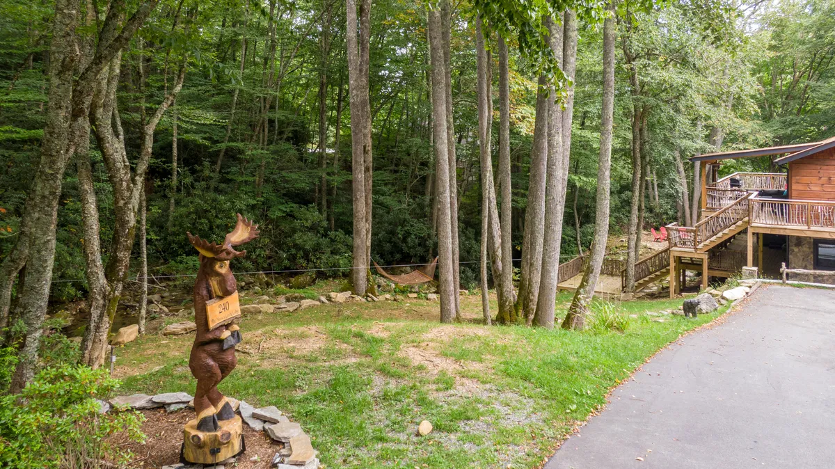 A tranquil view of the lodge’s exterior shows a log cabin-style house nestled among tall trees. A wooden moose statue adds a playful touch, while a hammock gently sways between the trees. The scene also features a cozy porch, inviting guests to relax and take in the natural surroundings.