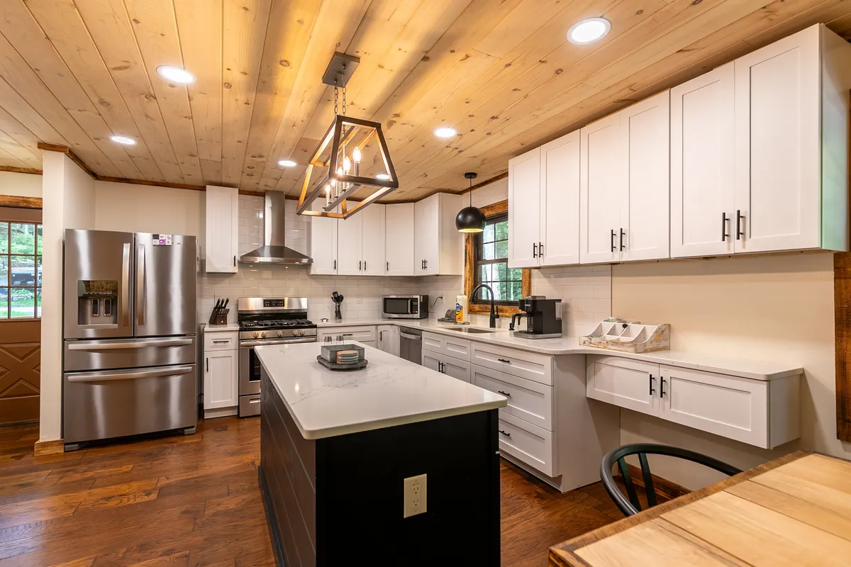 A bright, contemporary kitchen featuring a spacious island at the center, sleek modern appliances, warm wooden ceilings, and crisp white cabinetry. The design invites gathering and easy flow for both cooking and socializing, with ample workspace and gleaming surfaces.