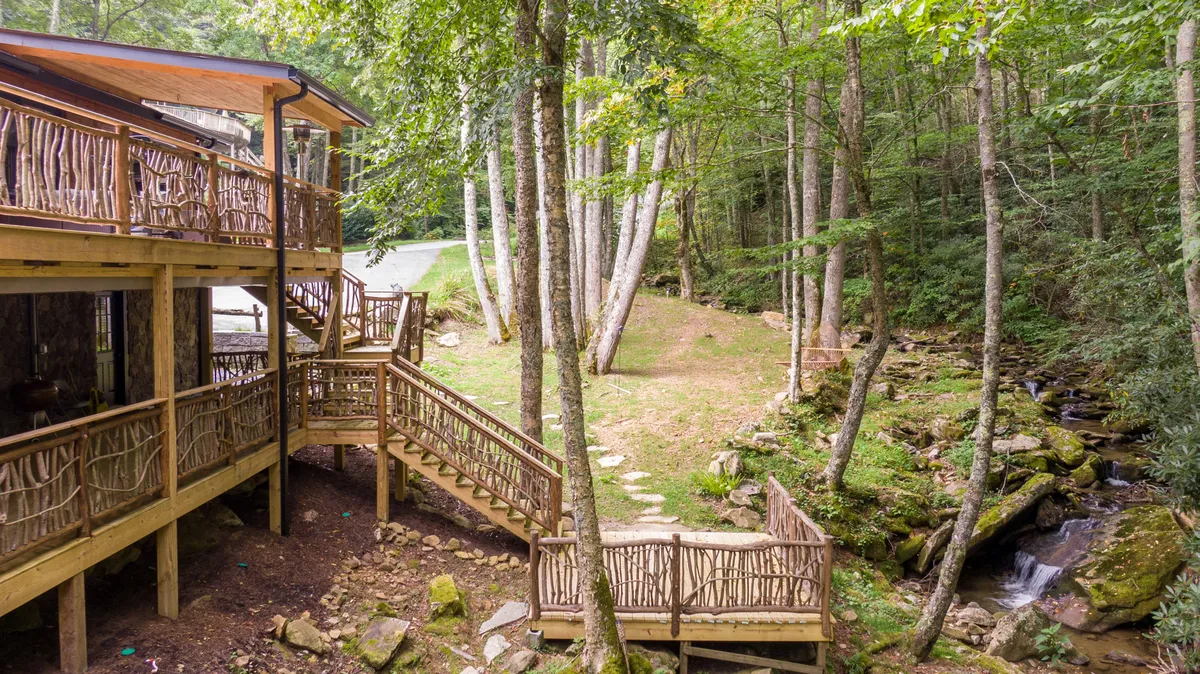 An inviting wooden deck set among tranquil trees with a stone pathway leading to a flowing creek. The deck is ideal for outdoor dining or lounging, offering peaceful views and the soothing sound of water nearby.