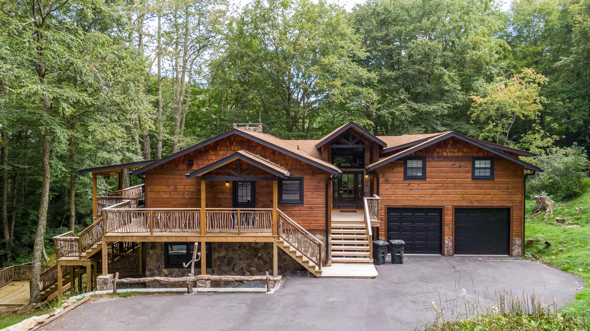 The image shows a stunning wooden lodge nestled among towering trees, with a spacious wraparound porch accented by stone details. The home exudes warmth and tranquility, set against a serene natural backdrop—perfect for relaxing outdoor gatherings as the sun sets.